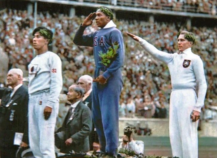Jesse Owens salutes during the presentation of his gold medal for the long jump, after defeating Germany’s Lutz Long during the 1936 Summer Olympics in Berlin (1936) [Colourised]